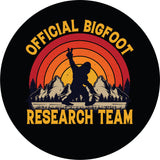 Sasquatch spare tire cover for Bronco, Jeep, RV, camper, and more with Bigfoot throwing out the peace sign as he walks along with mountains in the back and an ombre colored sunset. With the words, "official bigfoot research team"