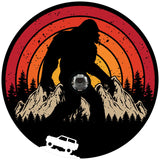 A retro vintage spare tire cover design with Sasquatch or bigfoot walking through the mountains and the silhouette of a Ford Bronco driving.  Designed for a black vinyl spare tire cover and a back up camera
